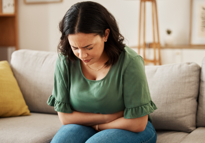 Frau sitzt auf der Couch, hat den Kopf gesenkt und hält sich mit verschränkten Armen den Bauch