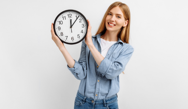 Junge Frau hält eine analoge Wanduhr neben sich und lächelt