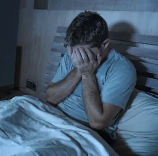 Mann sitzt verzweifelt nachts im Bett mit den Händen vor dem Gesicht