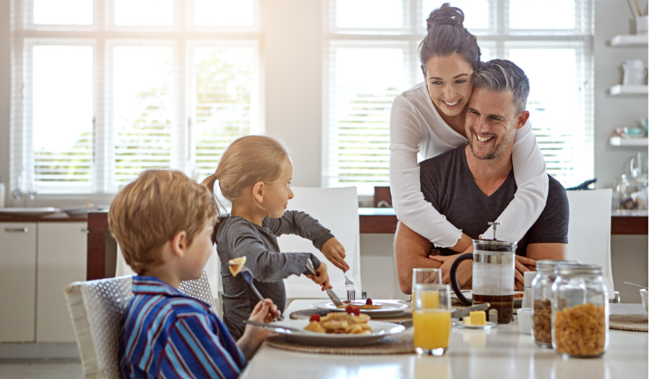Junge Familie beim Frühstücken am Esstisch. Mutter umarmt Vater. Kinder essen Pfannkuchen und trinken Orangensaft.